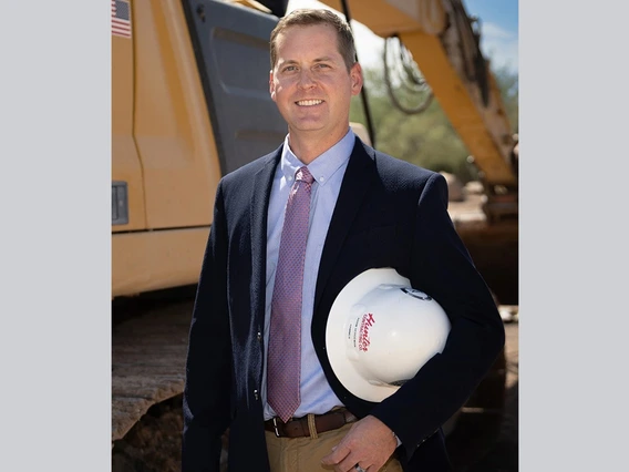 Man in coat in tie stands holding a hard hat in front of construction equipment.